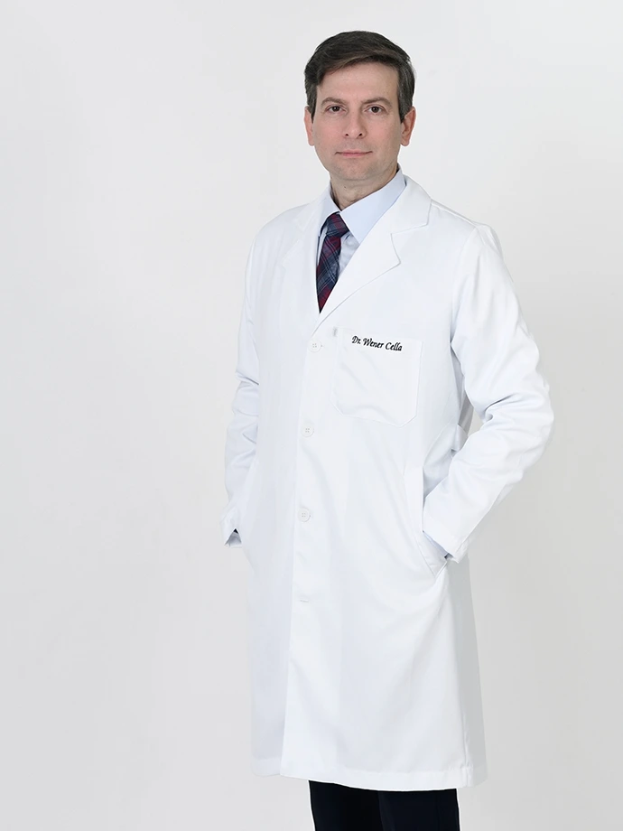 Retrato do Dr. Wener Cella, oftalmologista especialista em retina em São Luís, de jaleco branco com as mãos nos bolsos sobre fundo cinza