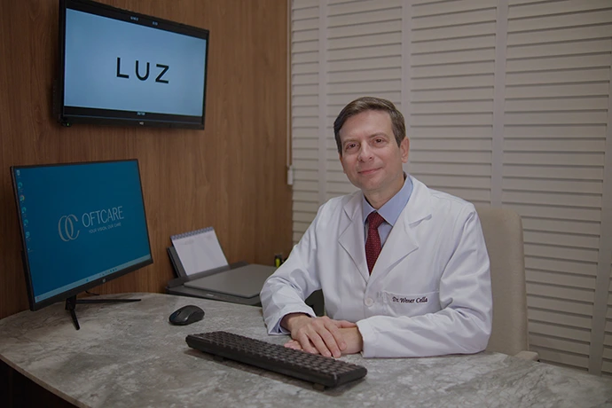 Dr. Wener Cella, oftalmologista de retina em São Luís, sentado em consultório com monitores exibindo LUZ e o logotipo OFTCARE