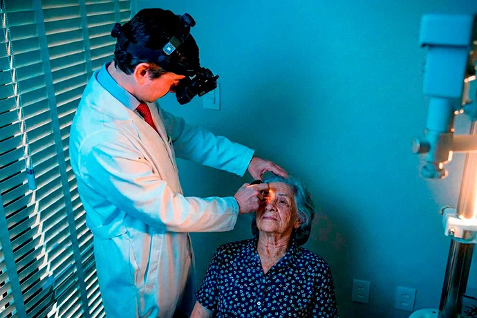 Oftalmologista Dr. Wener Cella, de jaleco branco e com lanterna de cabeça, examina o olho de uma paciente idosa sob luz forte e fria