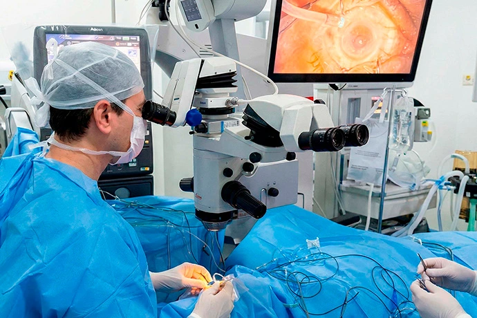 Oftalmologista Dr. Wener Cella, de uniforme azul, usando um microscópio cirúrgico durante uma operação, com uma visão ampliada do procedimento exibida em um monitor