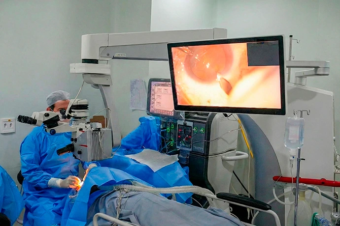 Oftalmologista Dr. Wener Cella, de uniforme azul, realiza um procedimento utilizando um microscópio cirúrgico enquanto um monitor exibe uma imagem ampliada do olho